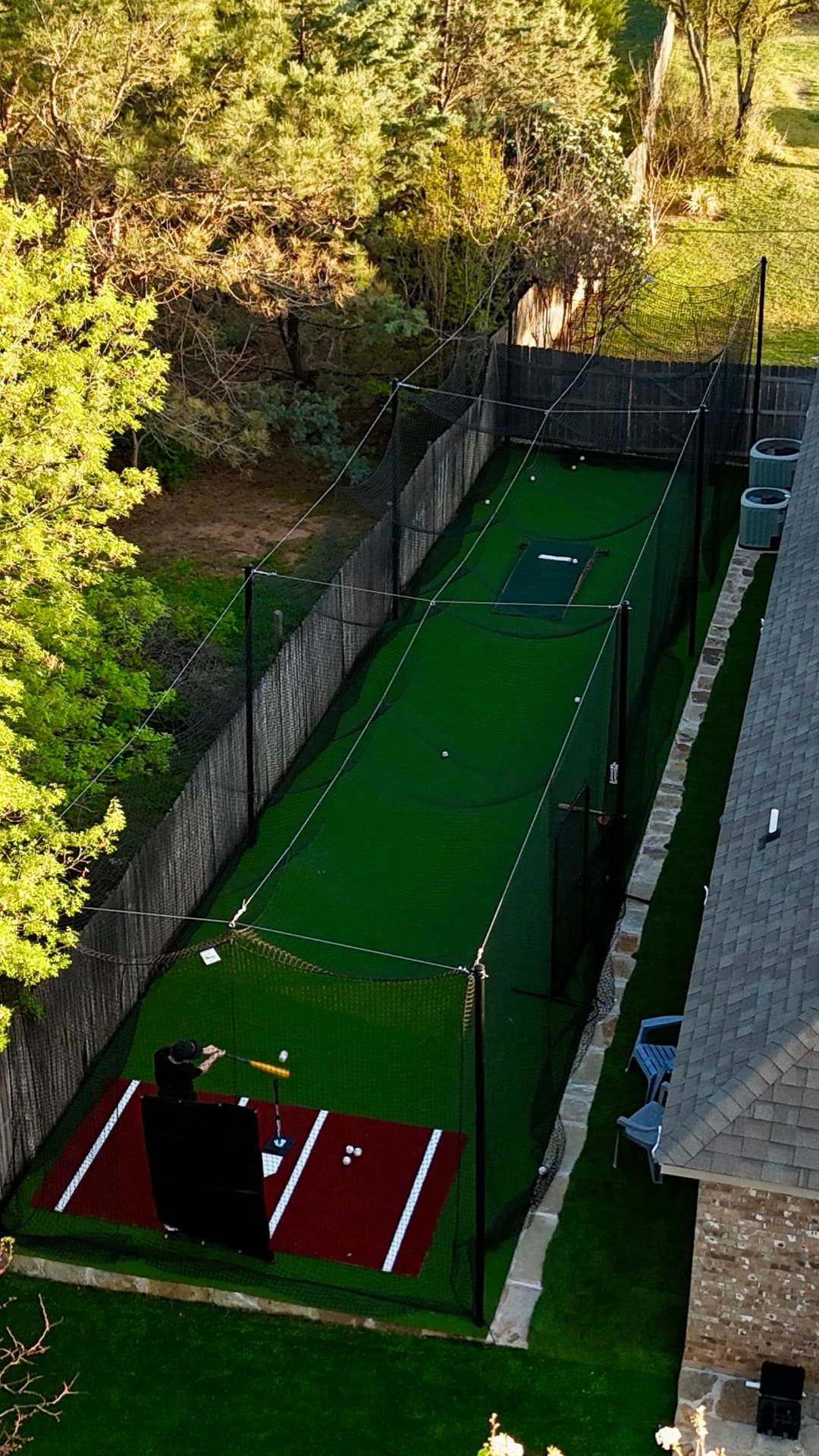 Aerial drone photo of backyard batting cage with artificial turf and black netting at golden hour beside stone home