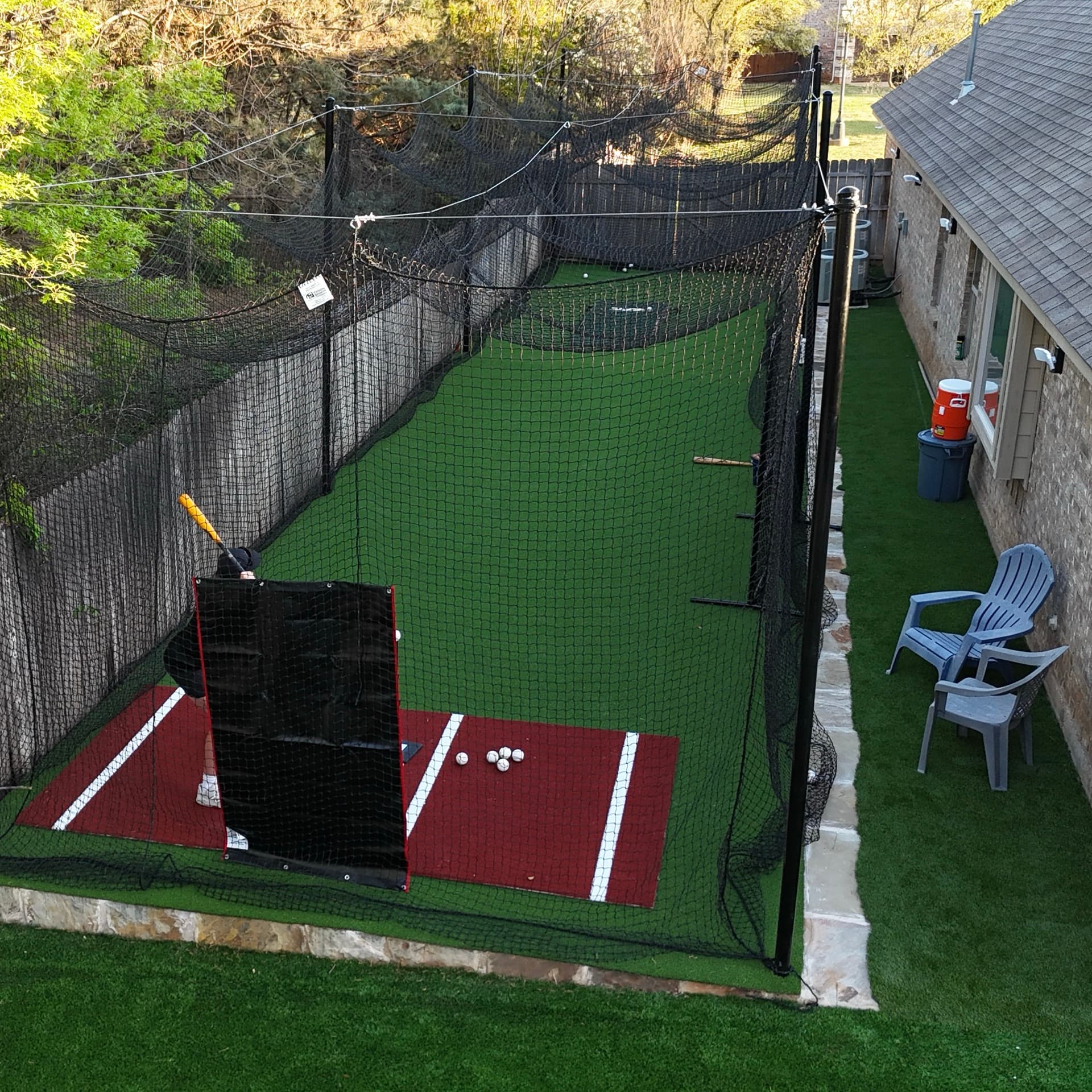 Overhead aerial of custom backyard batting cage with artificial turf, pitching machine, and netting beside residential home