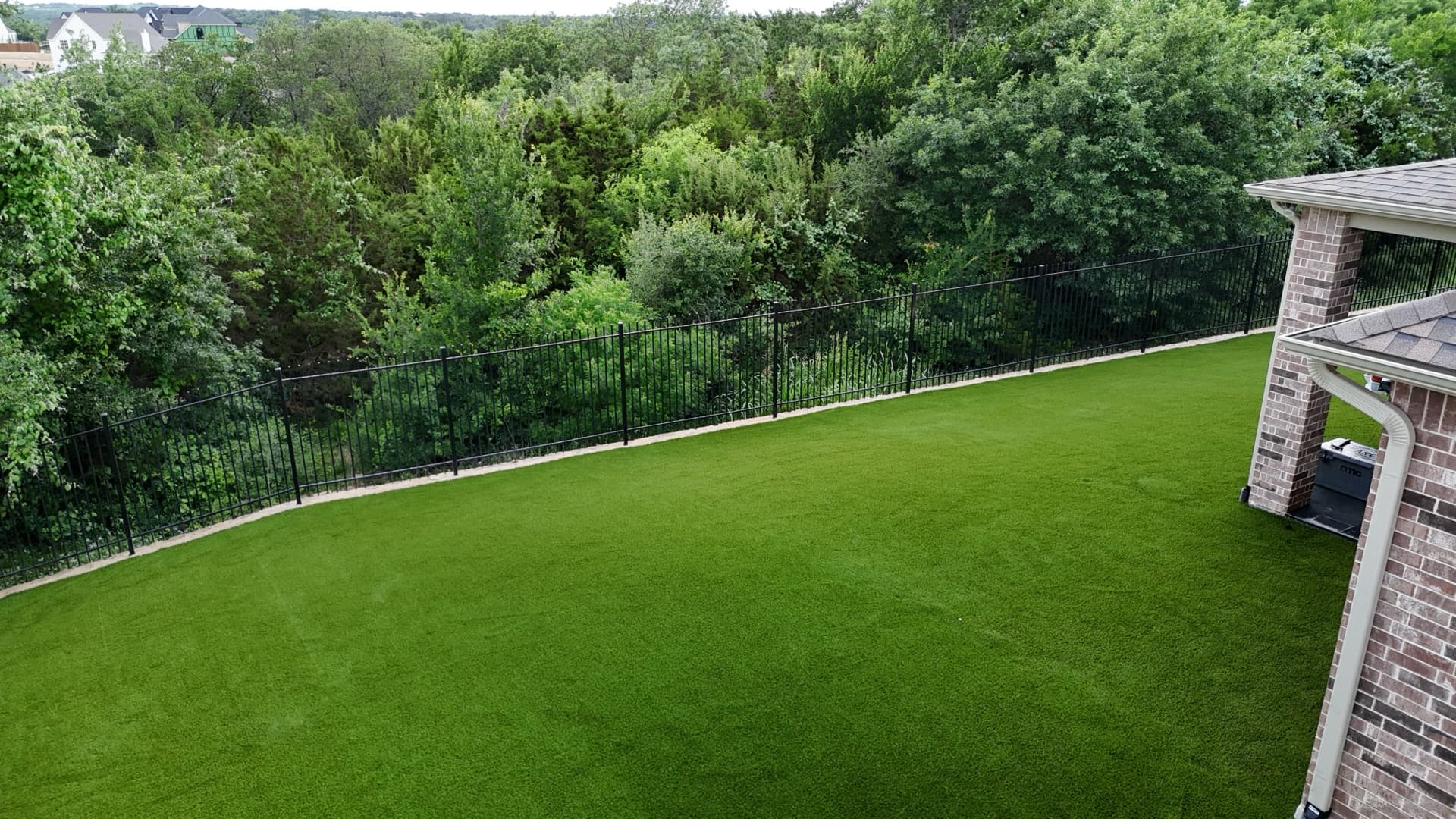 Aerial drone photo of completed backyard turf installation beside brick home with iron fence and cedar tree backdrop