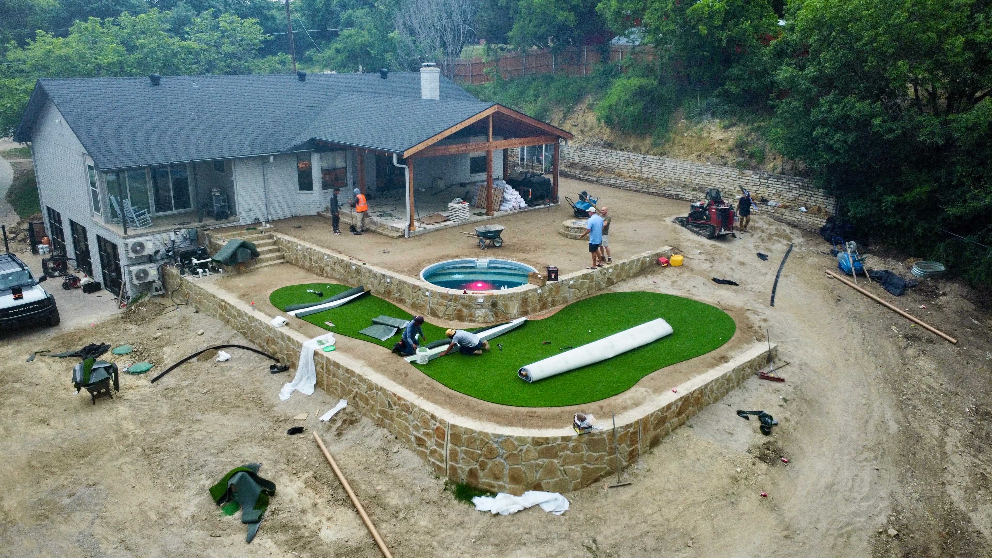 Aerial progress photo of backyard renovation showing crew installing artificial turf, plunge pool, and stone hardscape