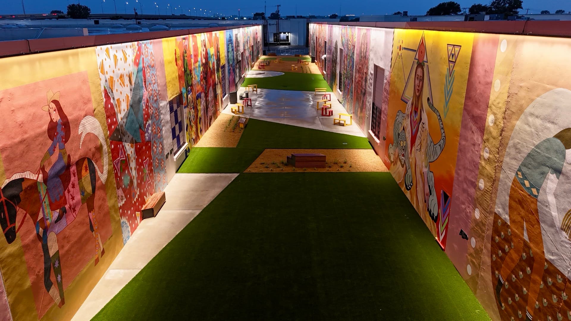 Ground-level aerial of the ArtDocks mural alley at twilight with glowing lights, vibrant murals, and deep blue evening sky