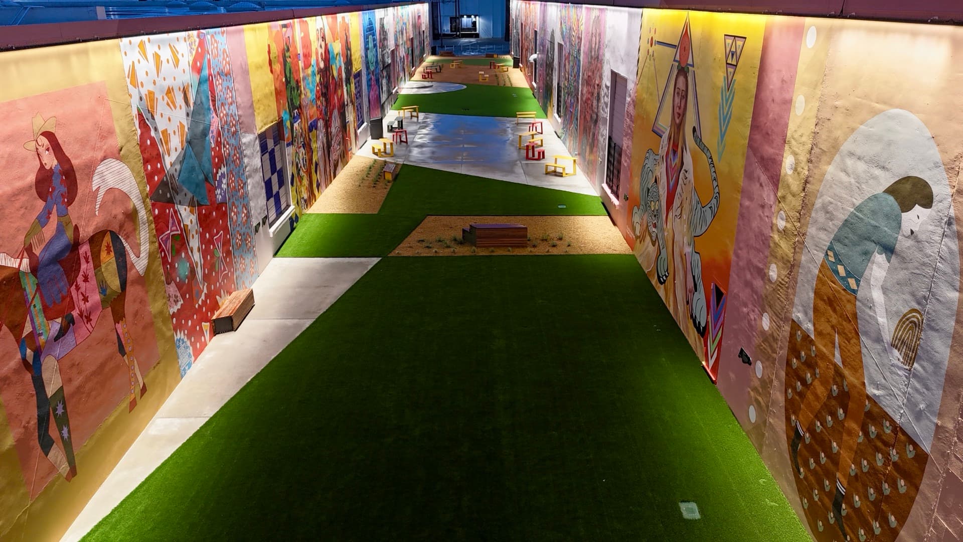 Wide aerial view looking down the ArtDocks mural alleyway with BearcatTurf artificial turf, colorful art murals, and seating at dusk