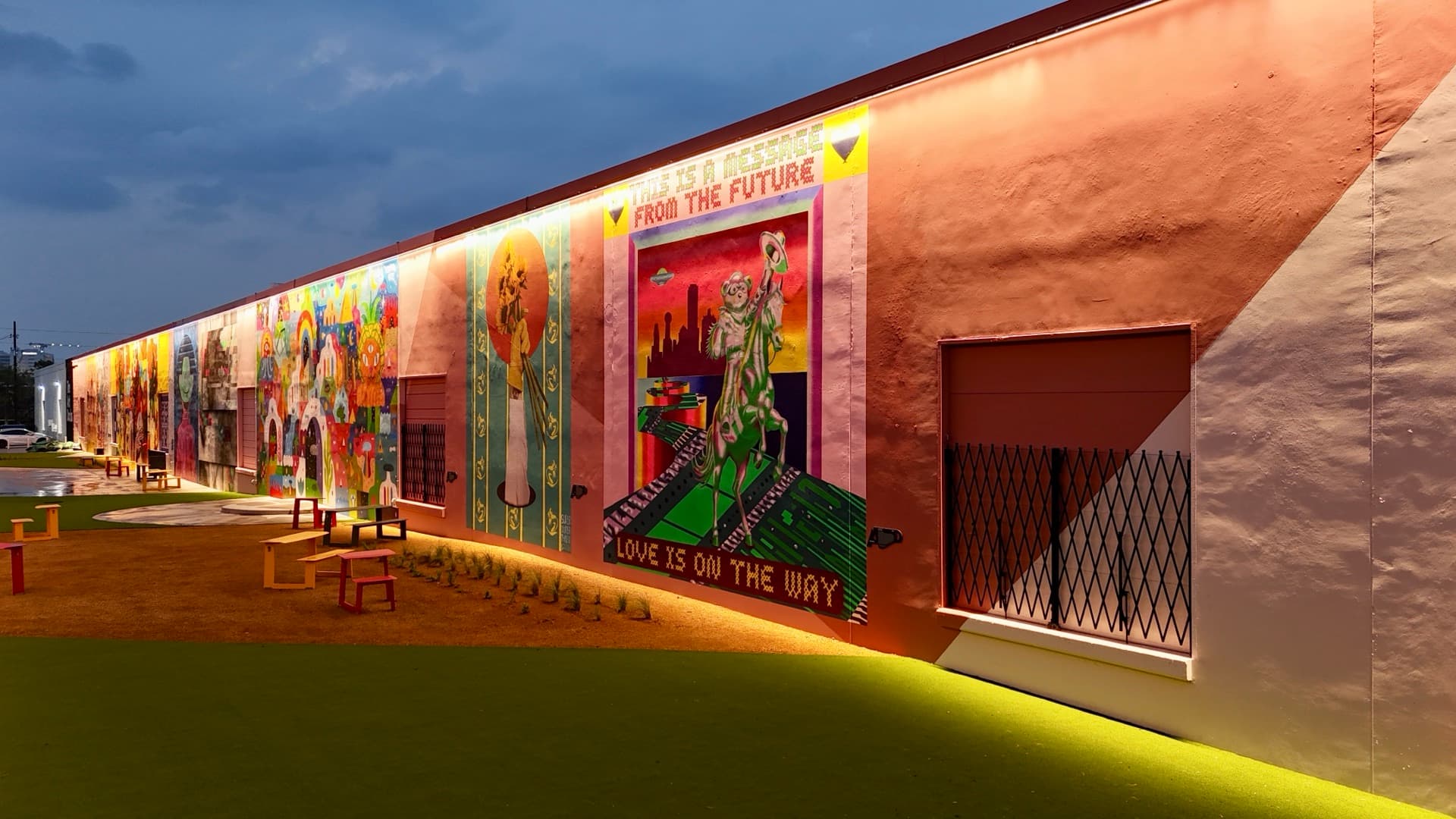 Aerial close-up of the ArtDocks mural wall at dusk showing vivid painted murals lit by ground-level uplighting