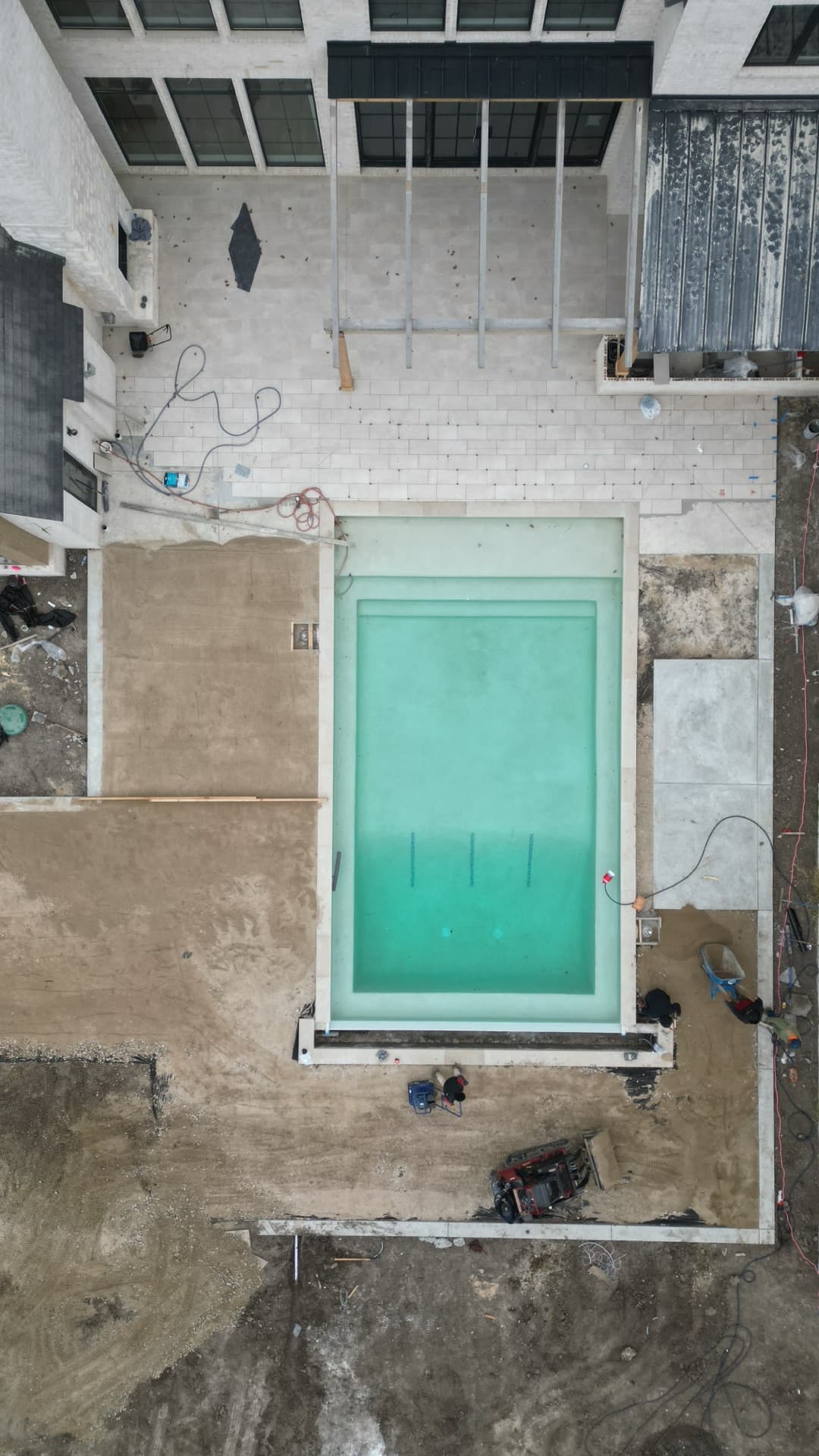 Straight-down overhead aerial of newly filled pool during luxury home construction with stone coping and surrounding dirt