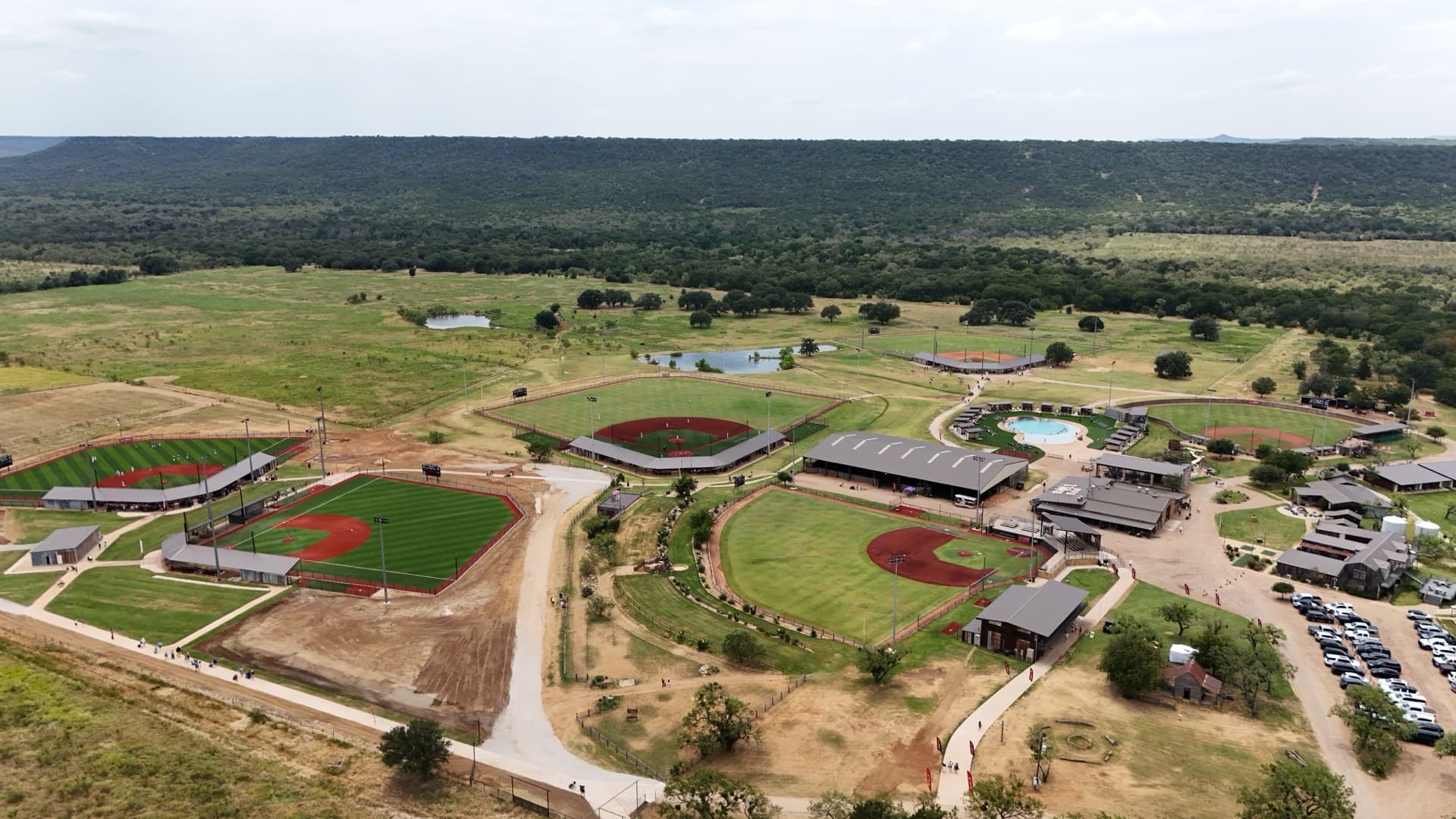 Aerial drone photo of Hill Country baseball resort showing four red-clay diamonds with grandstands, pond, and Palo Pinto mountains