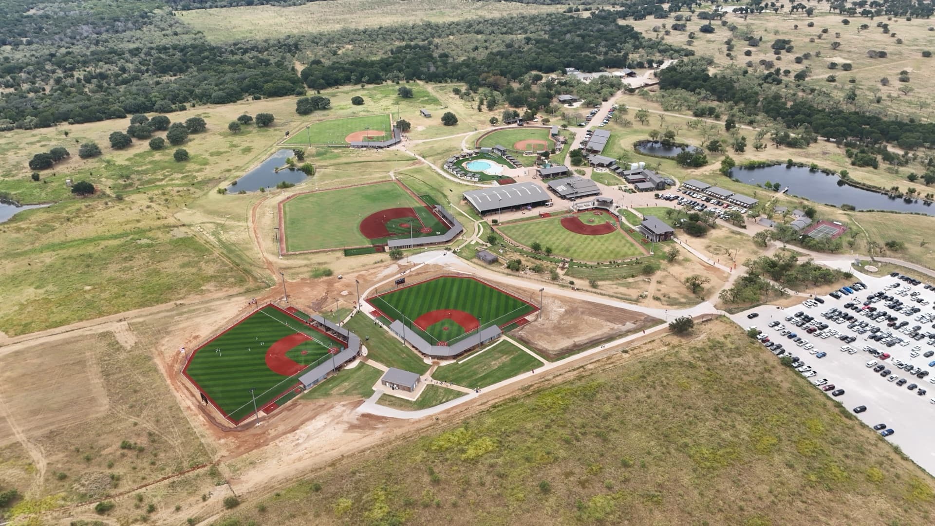 Wide aerial of Texas Hill Country baseball resort complex with four full diamonds, grandstands, pond, and cedar-covered hills