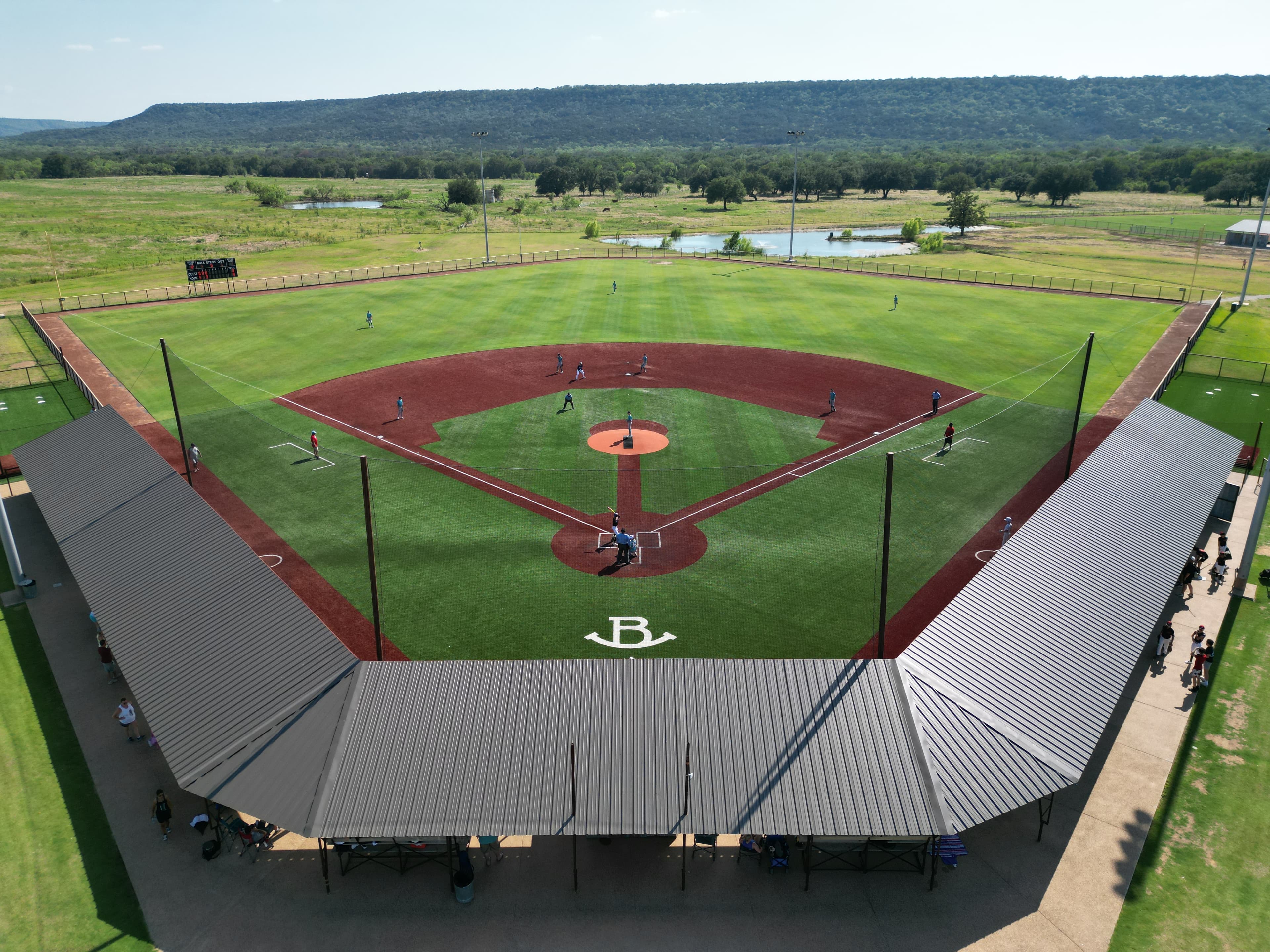 Aerial drone photo of baseball diamond with B logo, covered grandstands, players on field, and Hill Country landscape