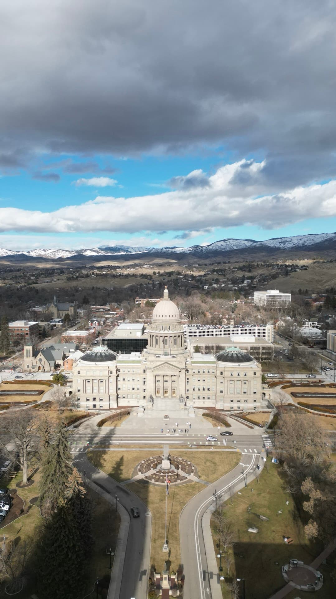 Vertical aerial of Idaho State Capitol with formal gardens, Boise cityscape, and snow-capped mountains beyond under dramatic clouds