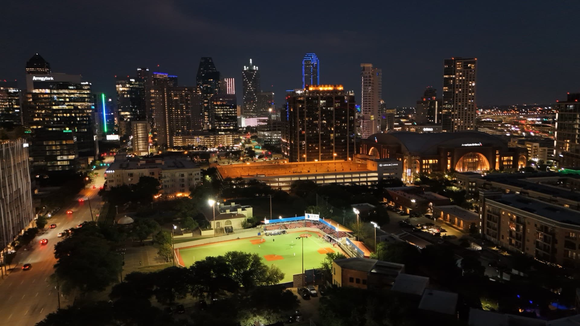Night aerial of Mavs Ballpark baseball field lit up with stadium lights and full downtown Dallas skyline illuminated behind