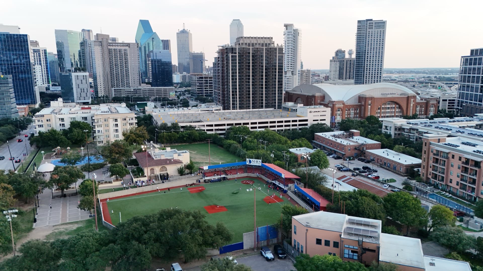 Aerial drone photo at dusk of Mavs Ballpark baseball field being set up with event crew visible and downtown Dallas skyline behind
