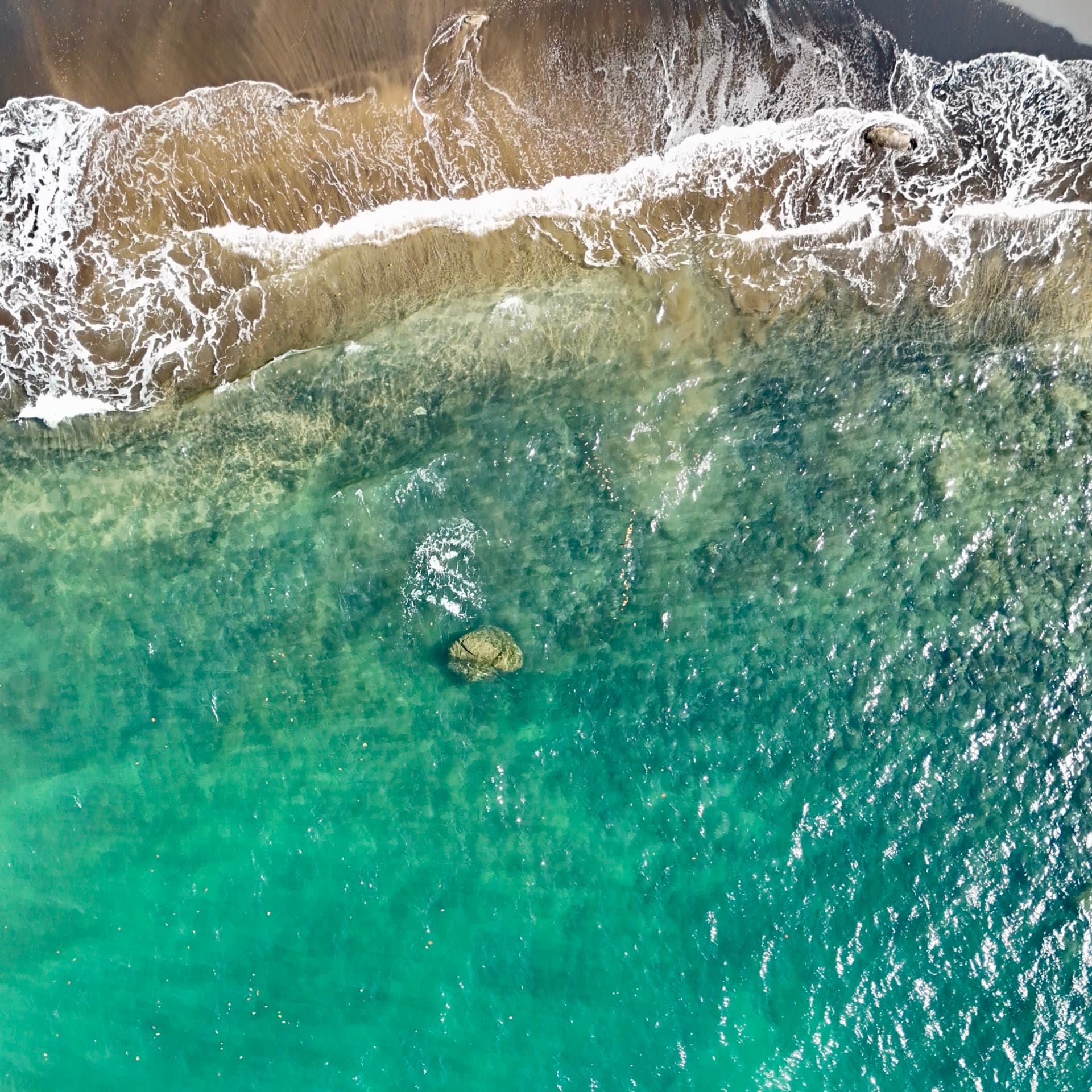 Overhead aerial drone photo of waves washing onto a dark sand beach with a single rock visible in clear shallow water