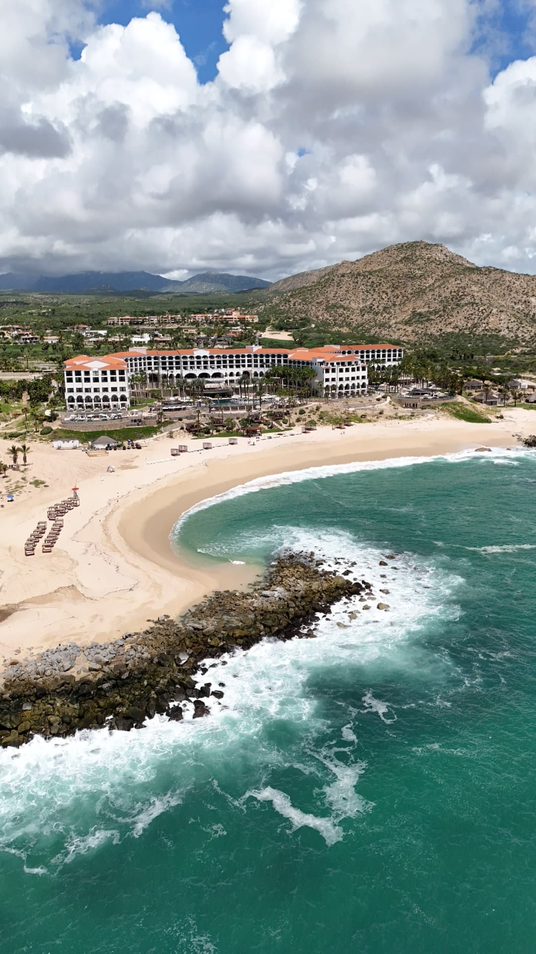 Vertical aerial of Cabo San Lucas beach peninsula with turquoise Pacific waves, resort hotels, and rocky coastline