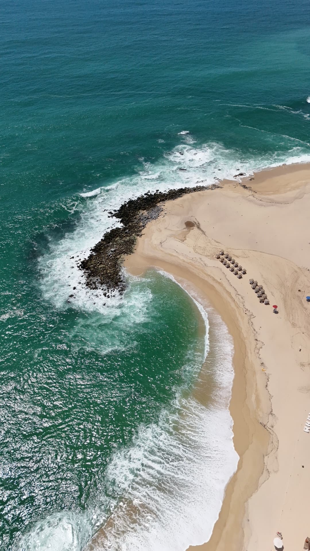 Overhead aerial of Cabo beach with turquoise breaking waves, white sand, and beach umbrella rows along shoreline