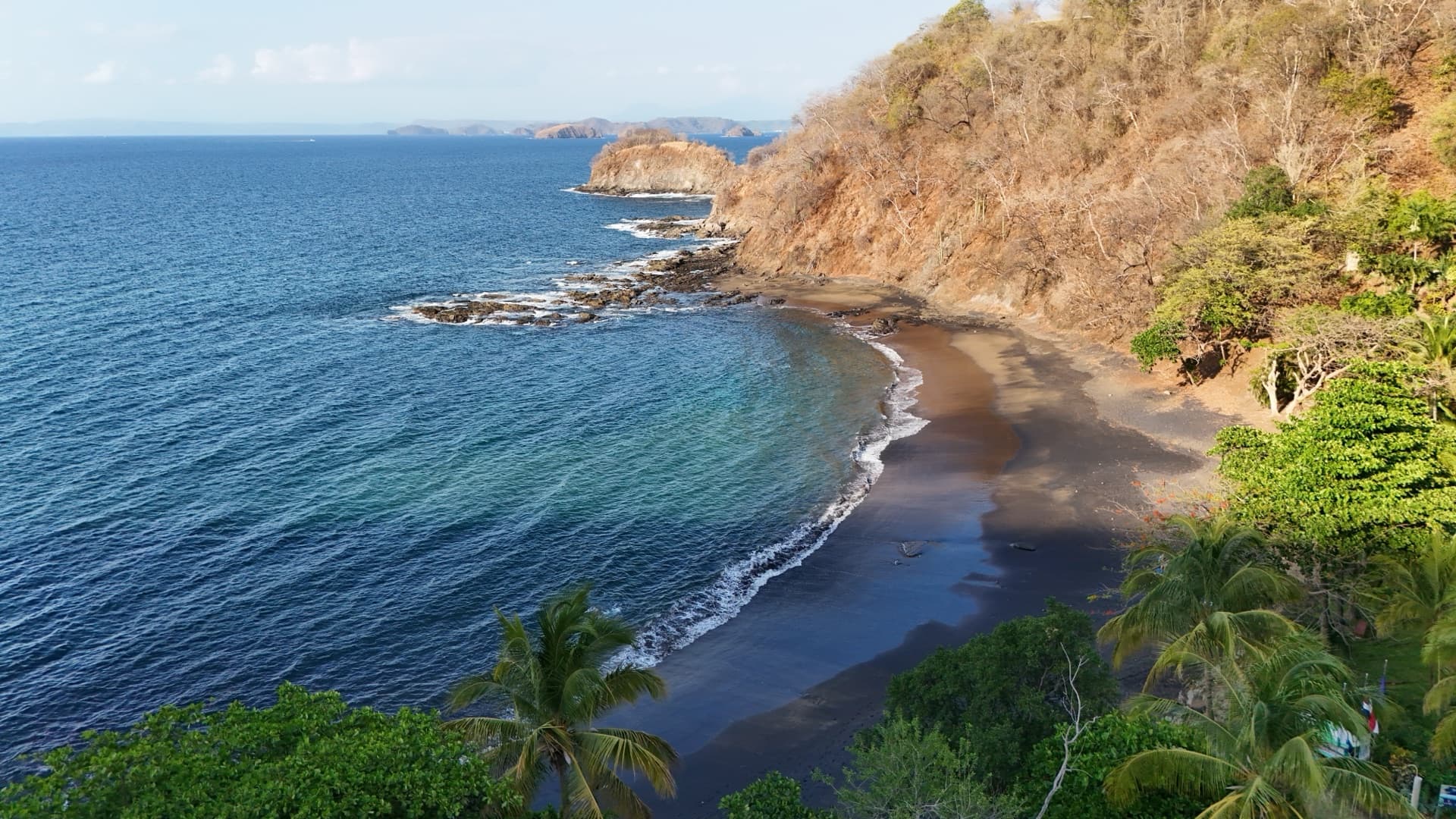 Aerial view of a dramatic dark sand beach cove with turquoise breaking waves, rocky coastline, and distant islands