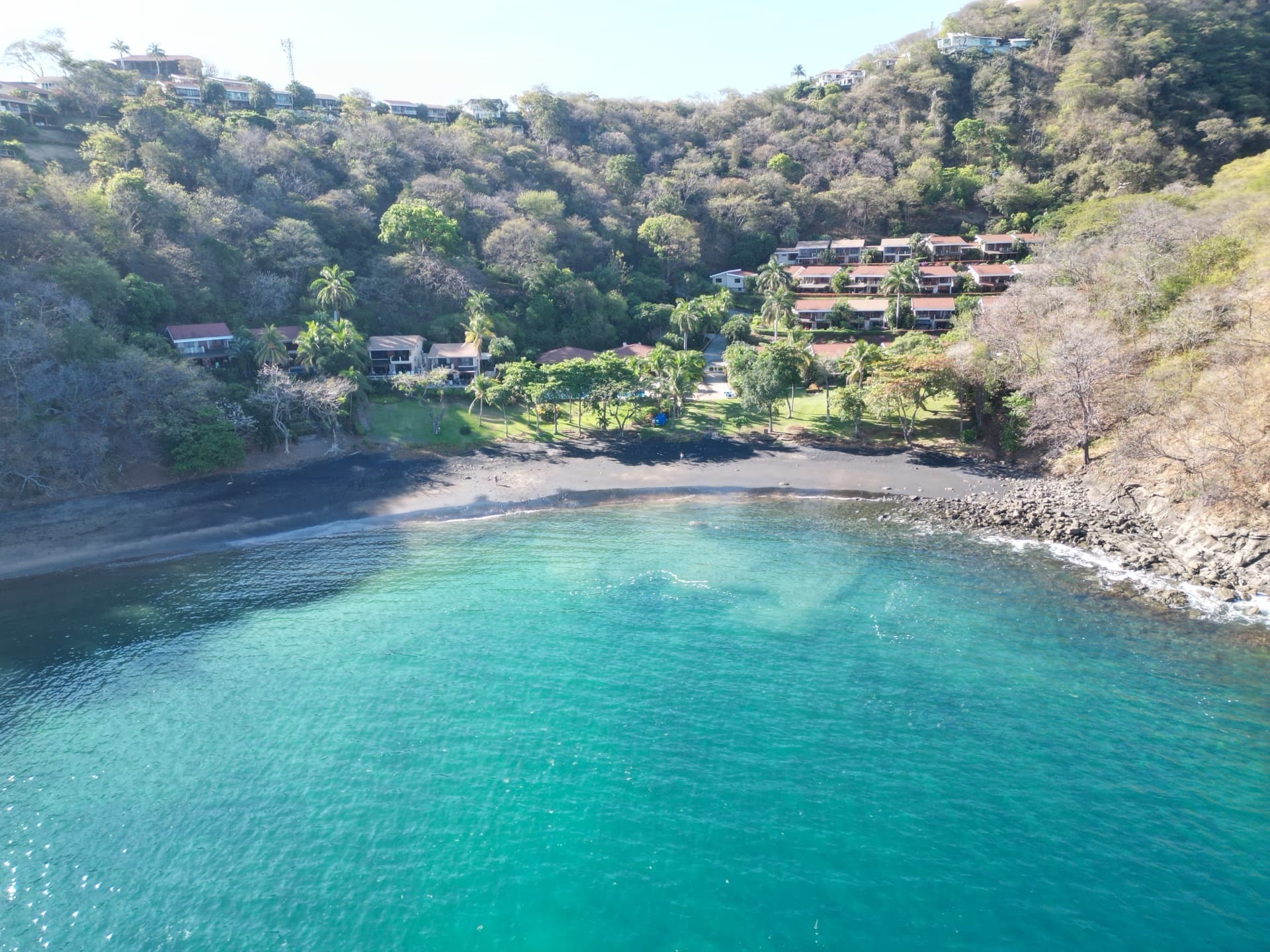 Low aerial of Costa Rica beach resort cove with turquoise water, dark sand beach, hillside villas, and tropical vegetation