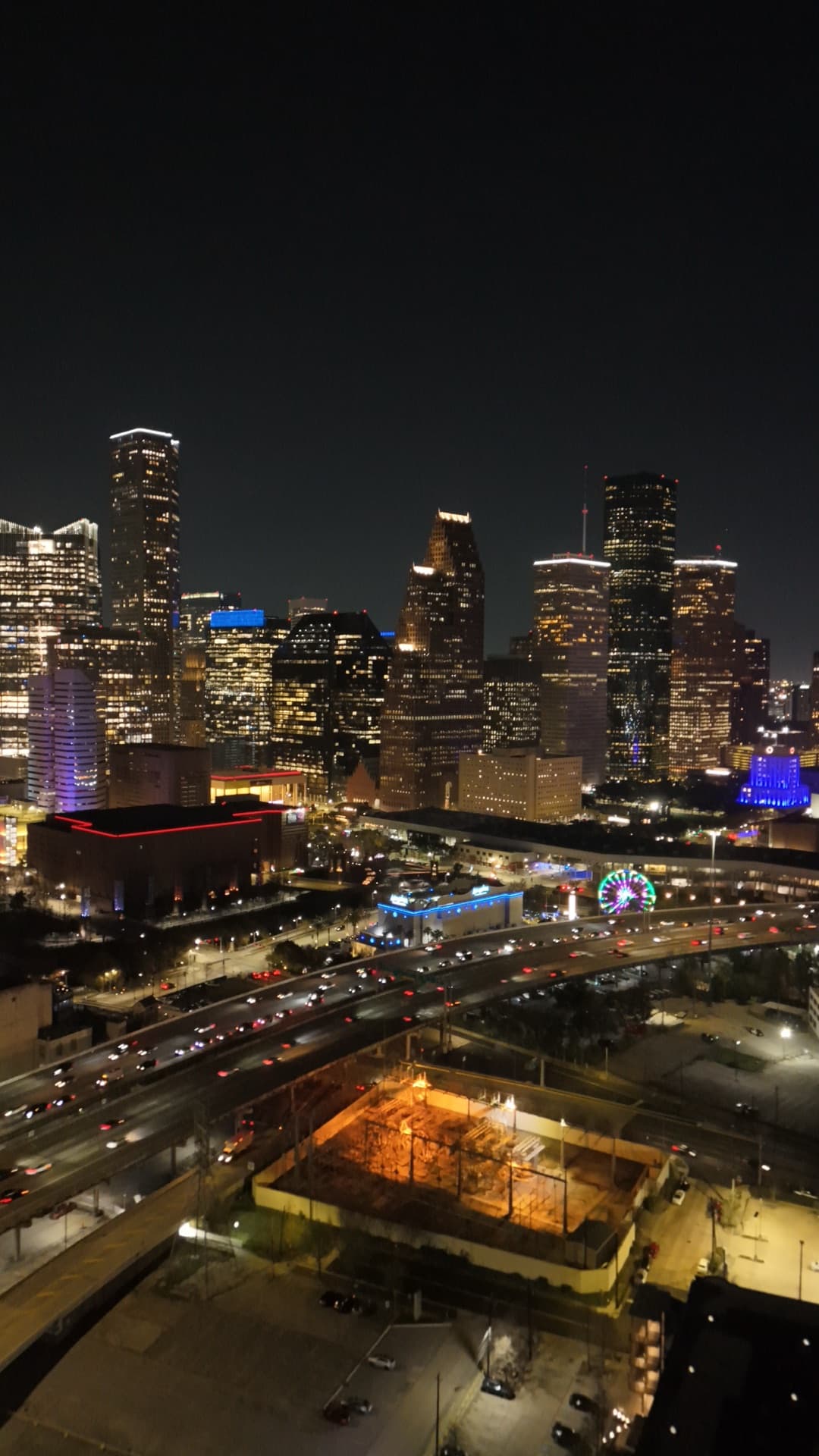 Vertical night aerial of downtown Houston skyline with glowing skyscrapers, highway interchange, and city lights