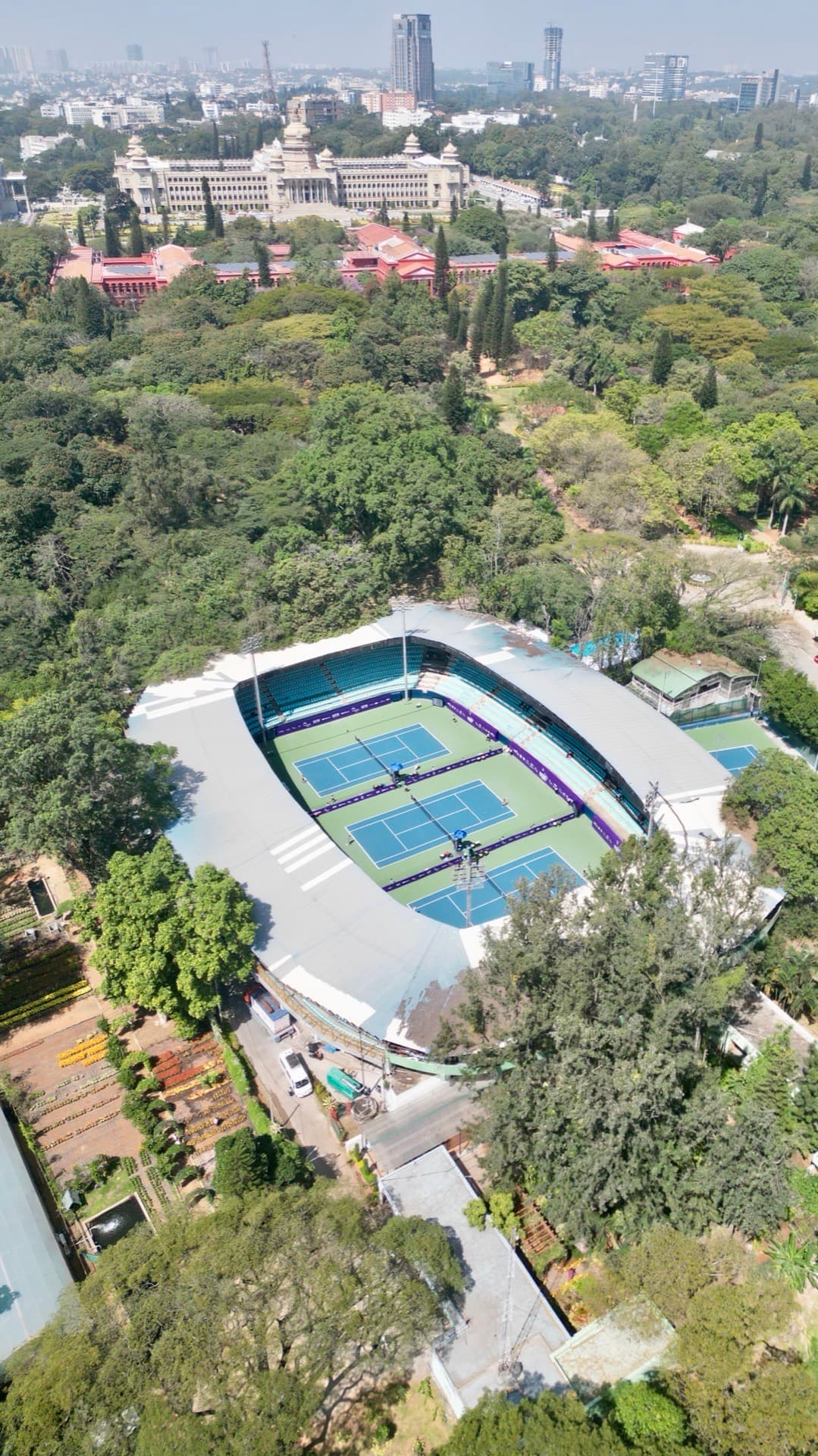 Aerial drone photo of a professional tennis stadium with blue courts, canopy roof, and surrounding tree canopy