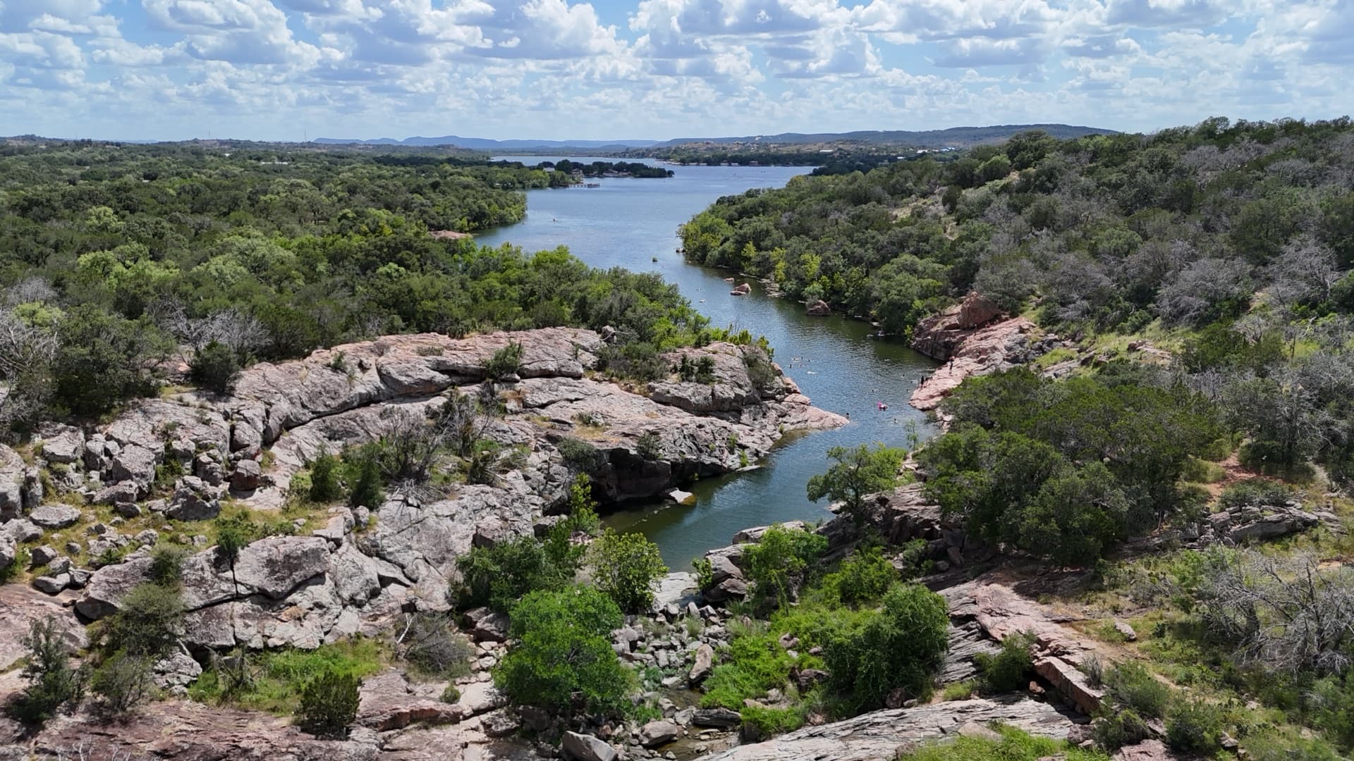 Wide aerial of Texas Hill Country lake with rugged limestone and granite canyon walls, cedar-covered bluffs, and open lake beyond