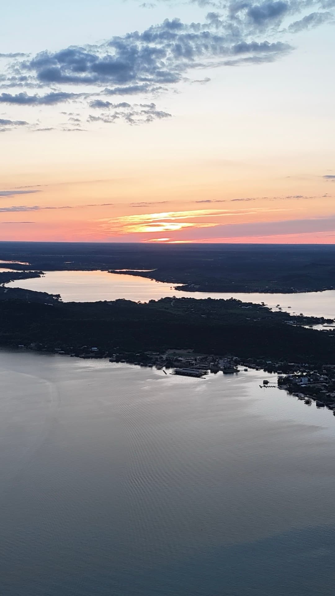 Vertical aerial at sunset over a wide Texas lake with glowing orange sky reflecting off calm water and wooded shoreline