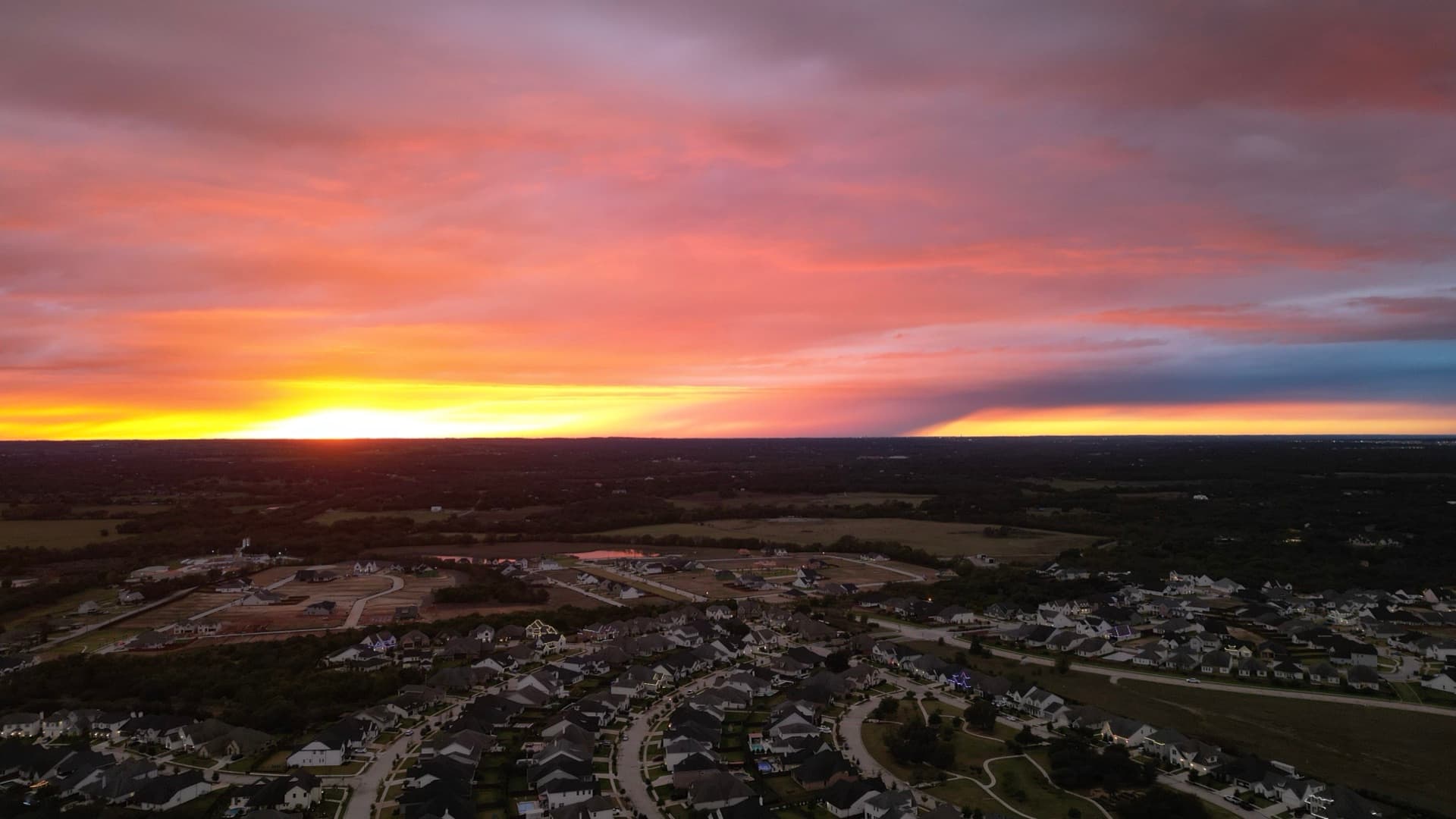 Dramatic wide aerial photo of a vivid red and orange Texas sunset over a suburban neighborhood with open fields