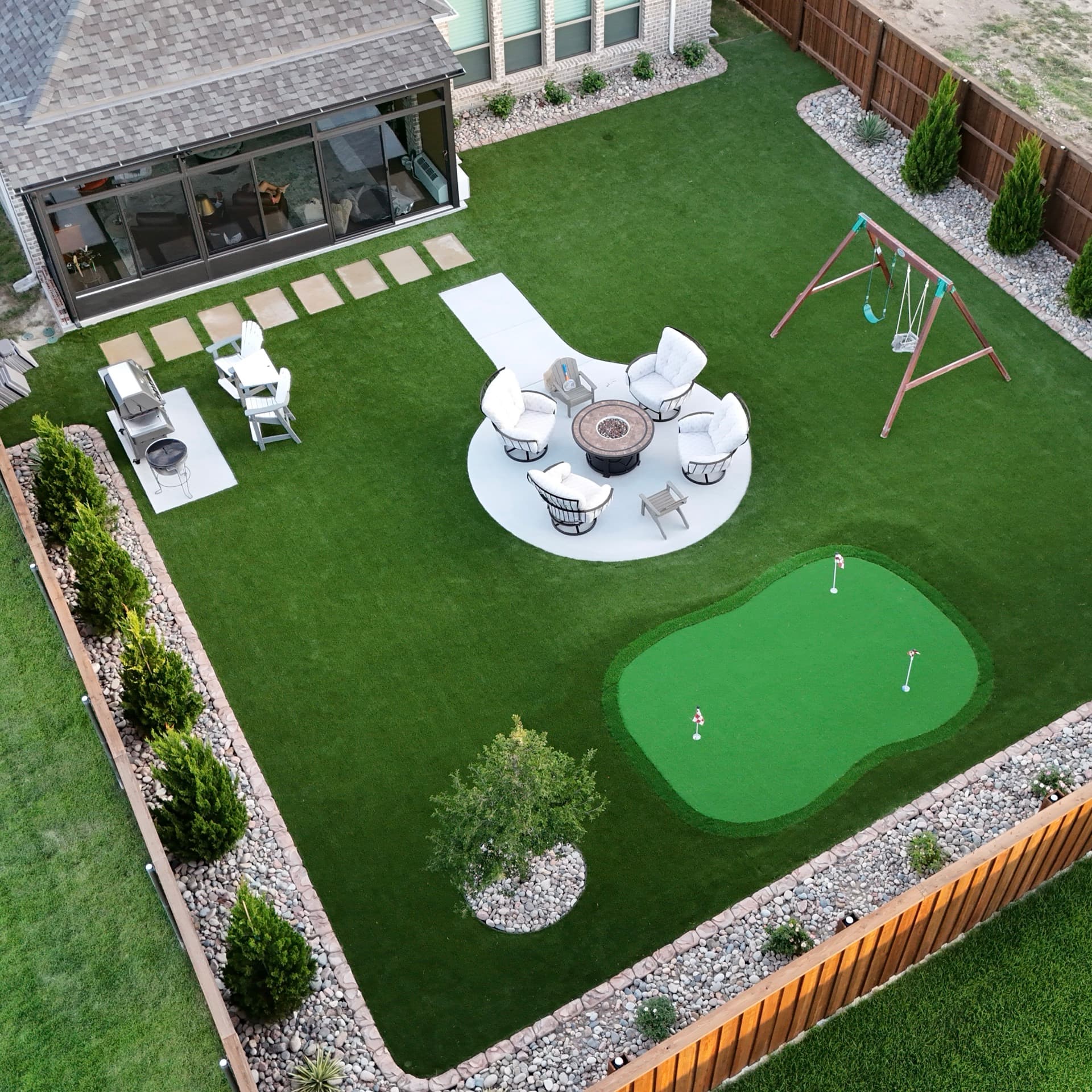 Overhead aerial of modern backyard with artificial turf, putting green, circular fire pit seating area, swing set, and decorative planters