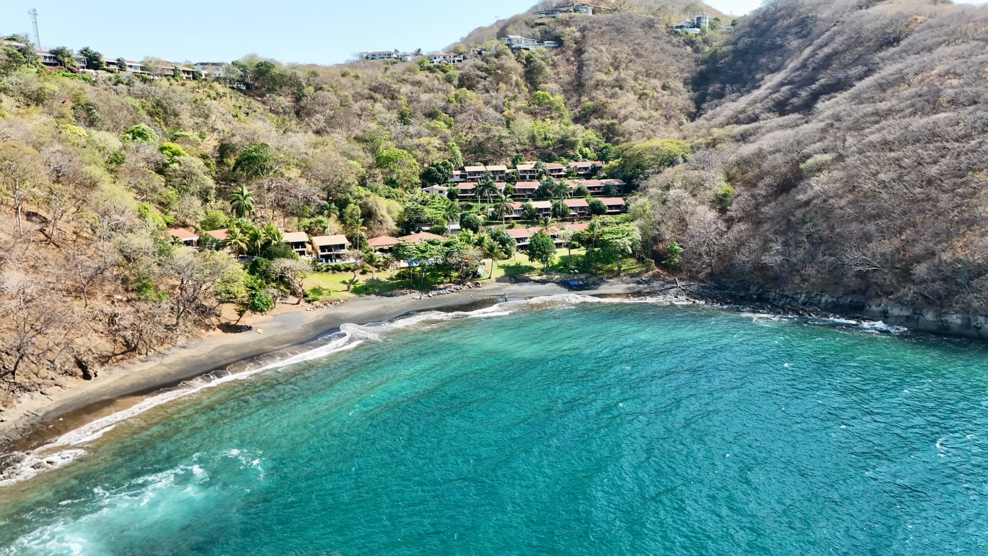 Aerial drone photo of a beach resort nestled in a dark sand cove with turquoise water, palm trees, and hillside villas