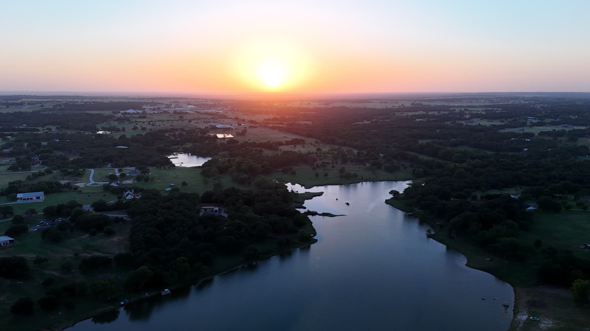 Golden hour aerial drone photo over a lake on a North Texas ranch property with warm light across the water and tree canopy