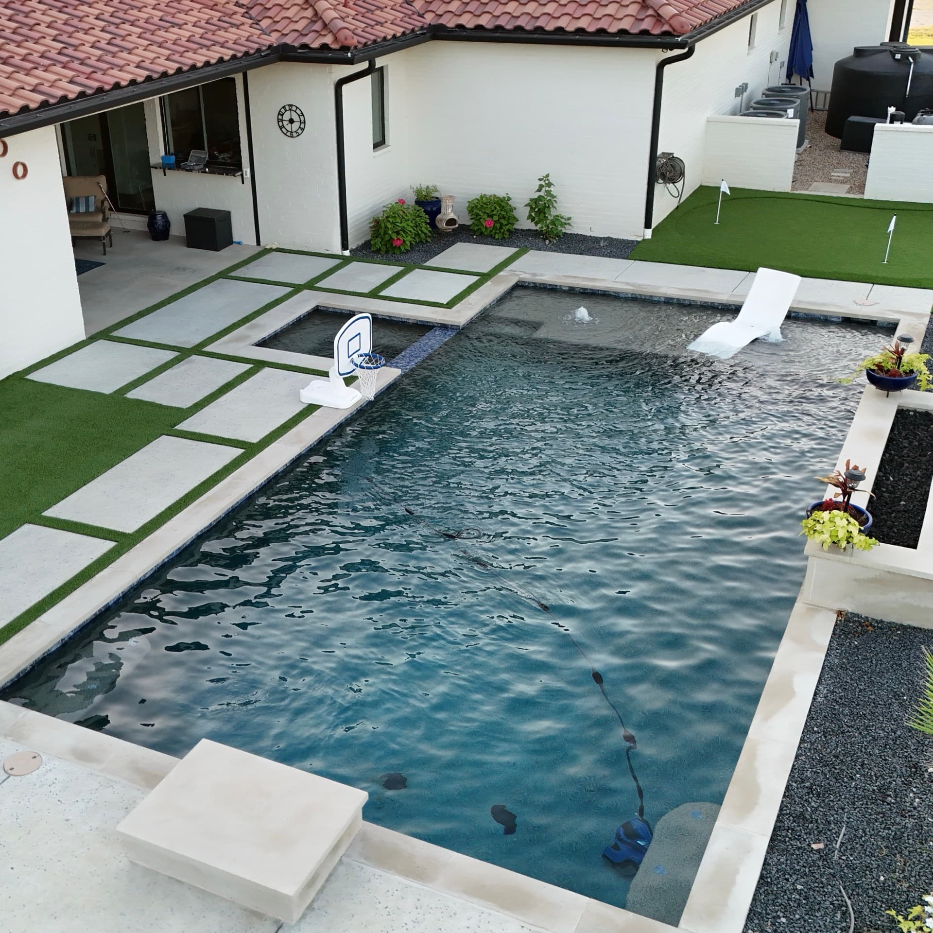 Aerial drone photo of luxury home backyard with rectangular pool, basketball hoop float, covered patio, and artificial turf lawn
