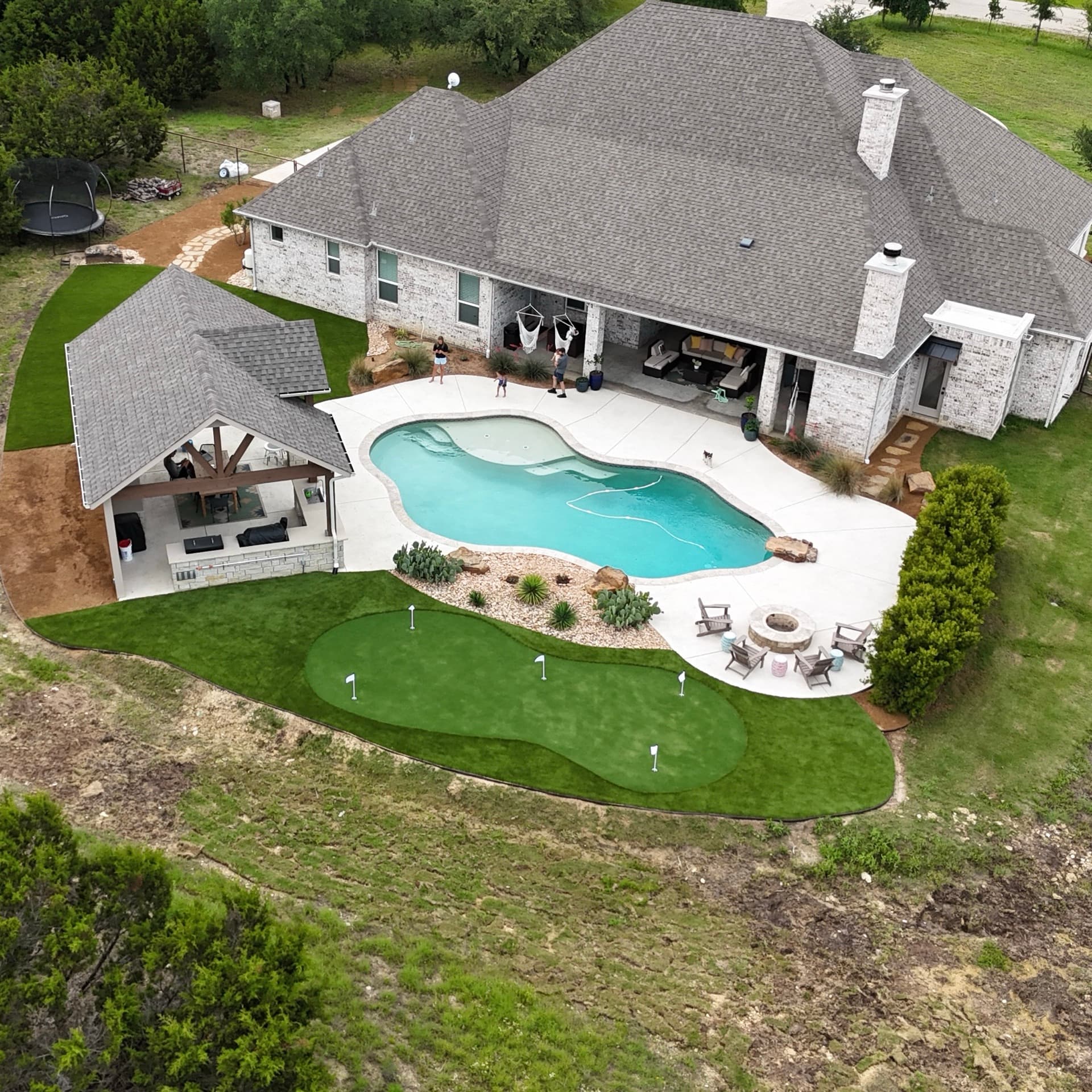 Aerial drone photo of Texas Hill Country home with freeform pool, covered patio, putting green, and fire pit in manicured backyard