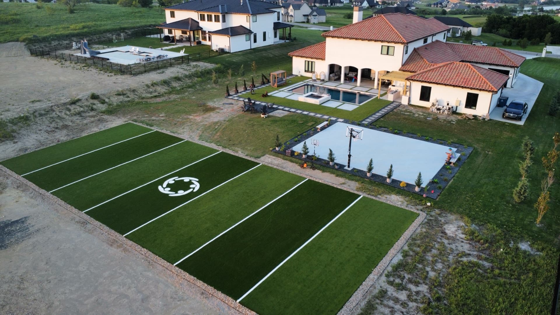 Wide aerial of luxury estate with soccer turf field, basketball court, and pool against open Texas landscape at dusk