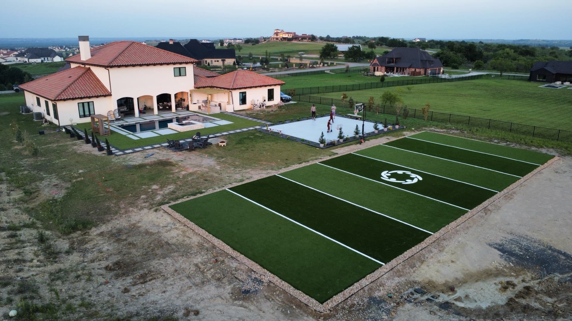 Aerial of luxury estate turf soccer field and basketball court with people playing and pool visible beside red-tile home