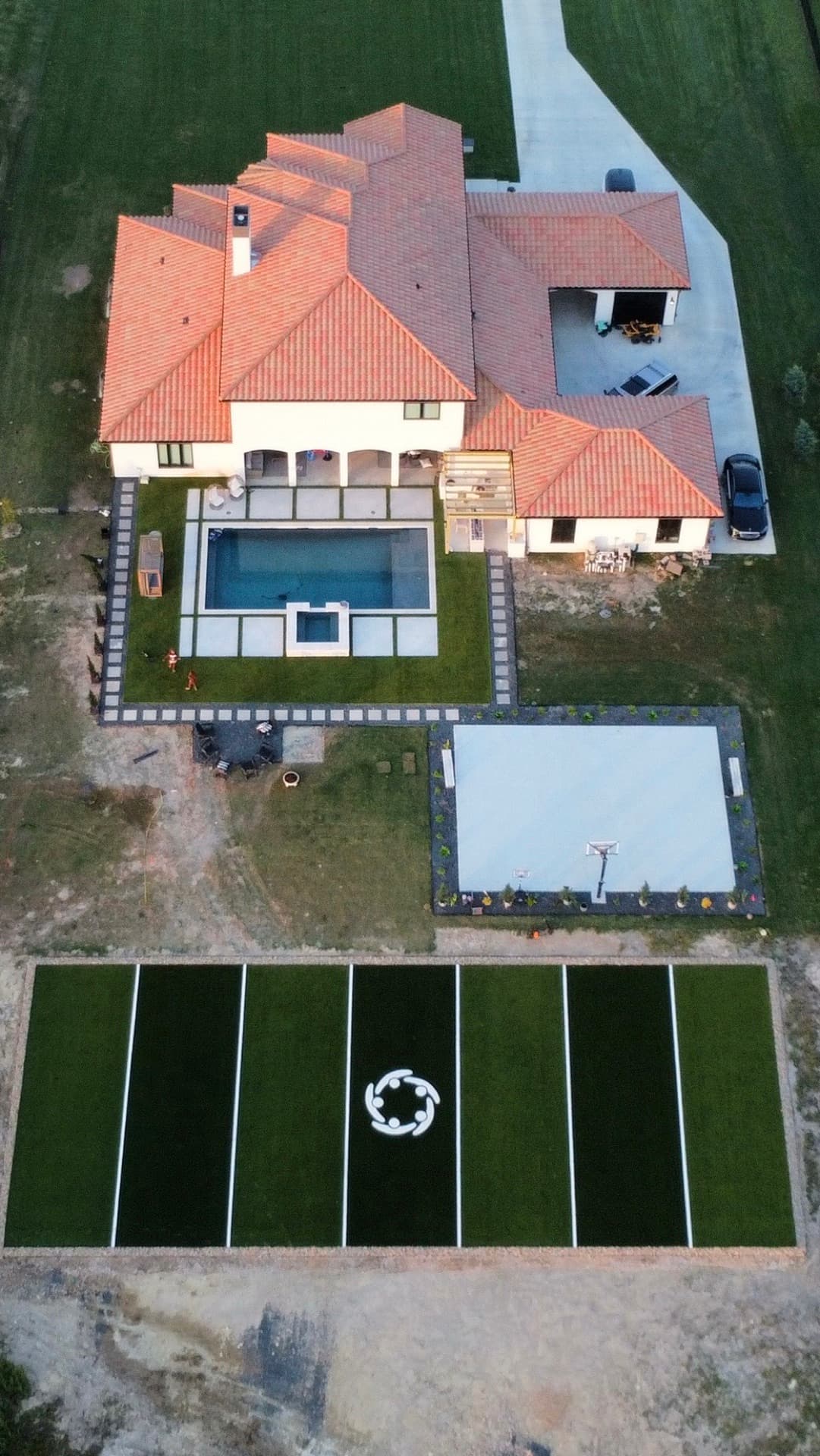 Overhead aerial of Spanish-style luxury estate with full soccer turf field, basketball court, and pool with spa at dusk