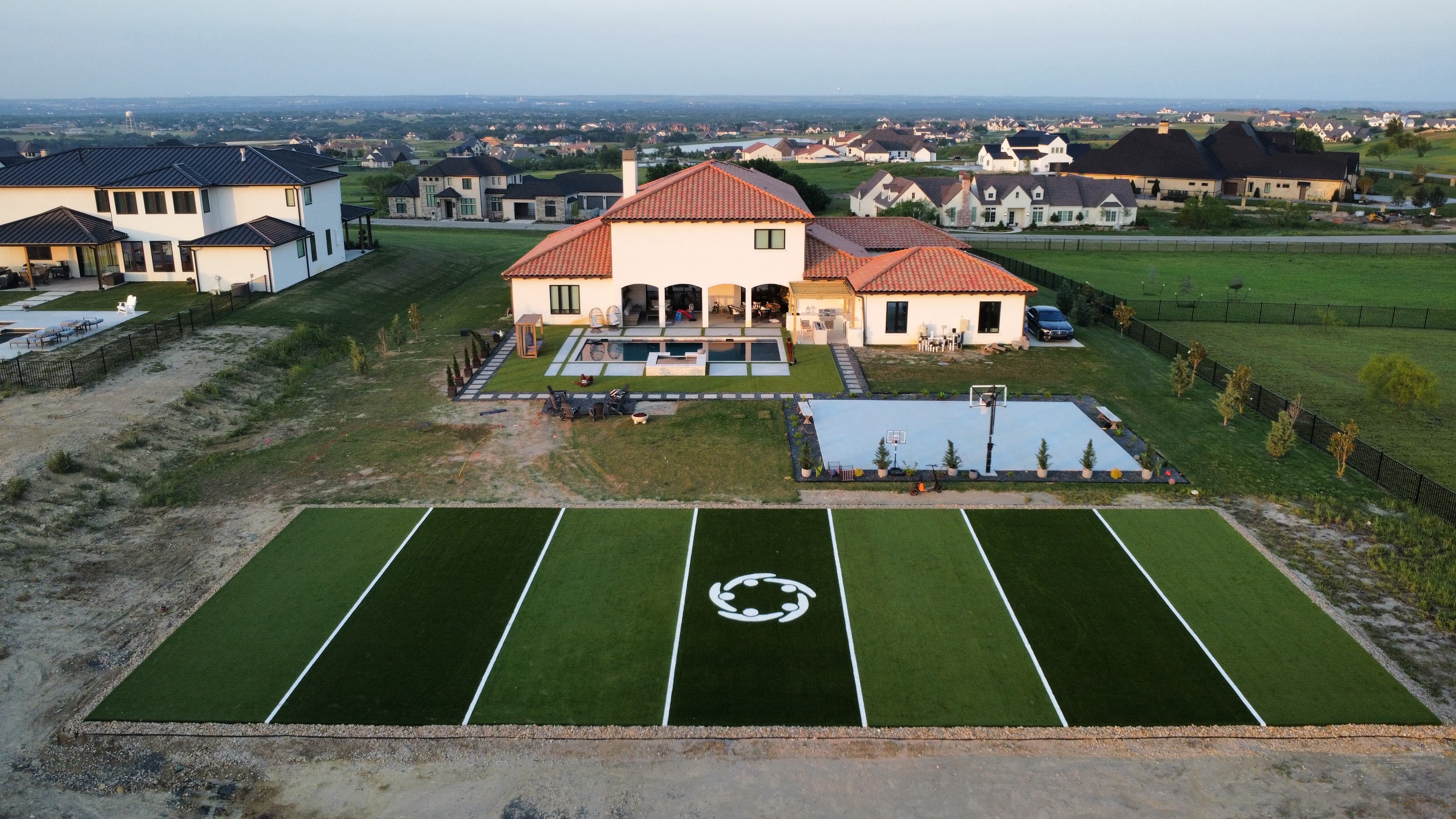 Aerial view of luxury home with private soccer field, pool, basketball court, and red tile roof at dusk