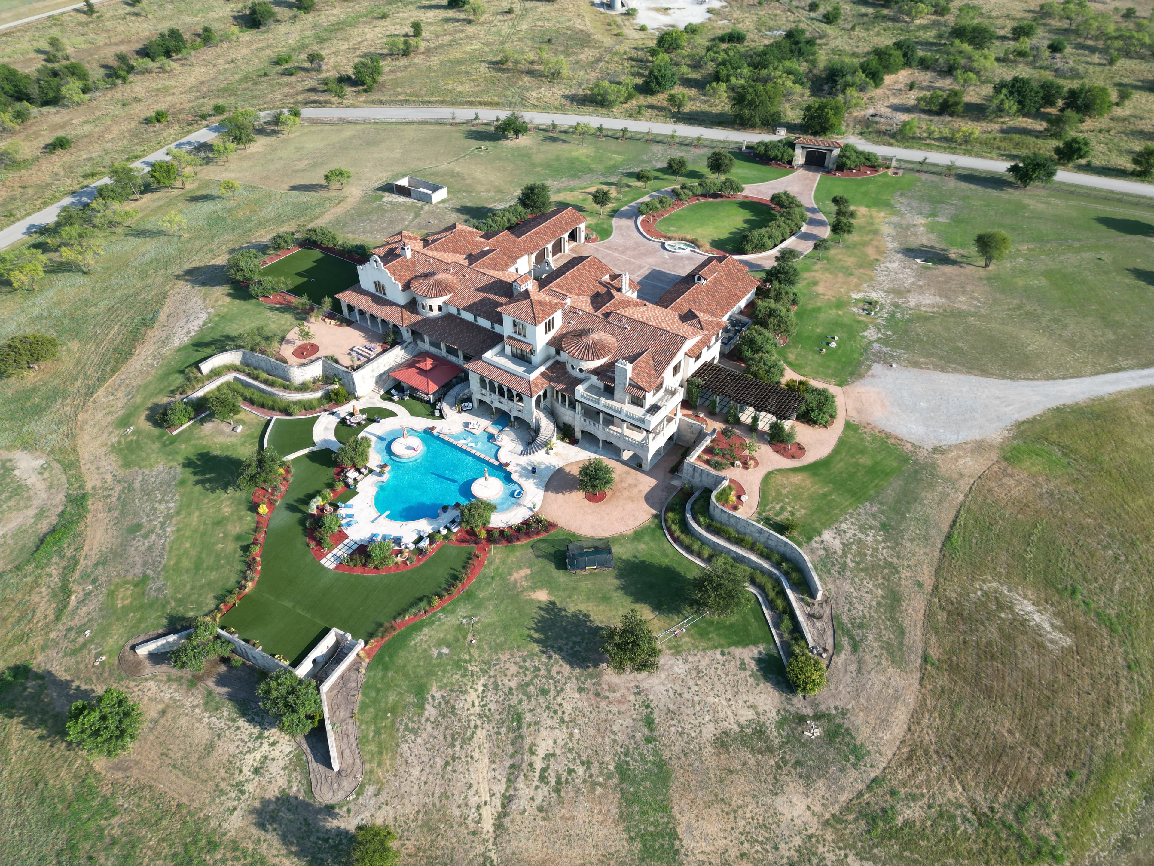 Aerial view of luxury Mediterranean estate with large pool, manicured grounds, and red tile roof in Texas Hill Country